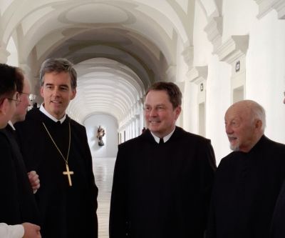 Mönche in schwarzem Ordengewand begrüßen einen Gast, im Hintergrund ein langer Gang